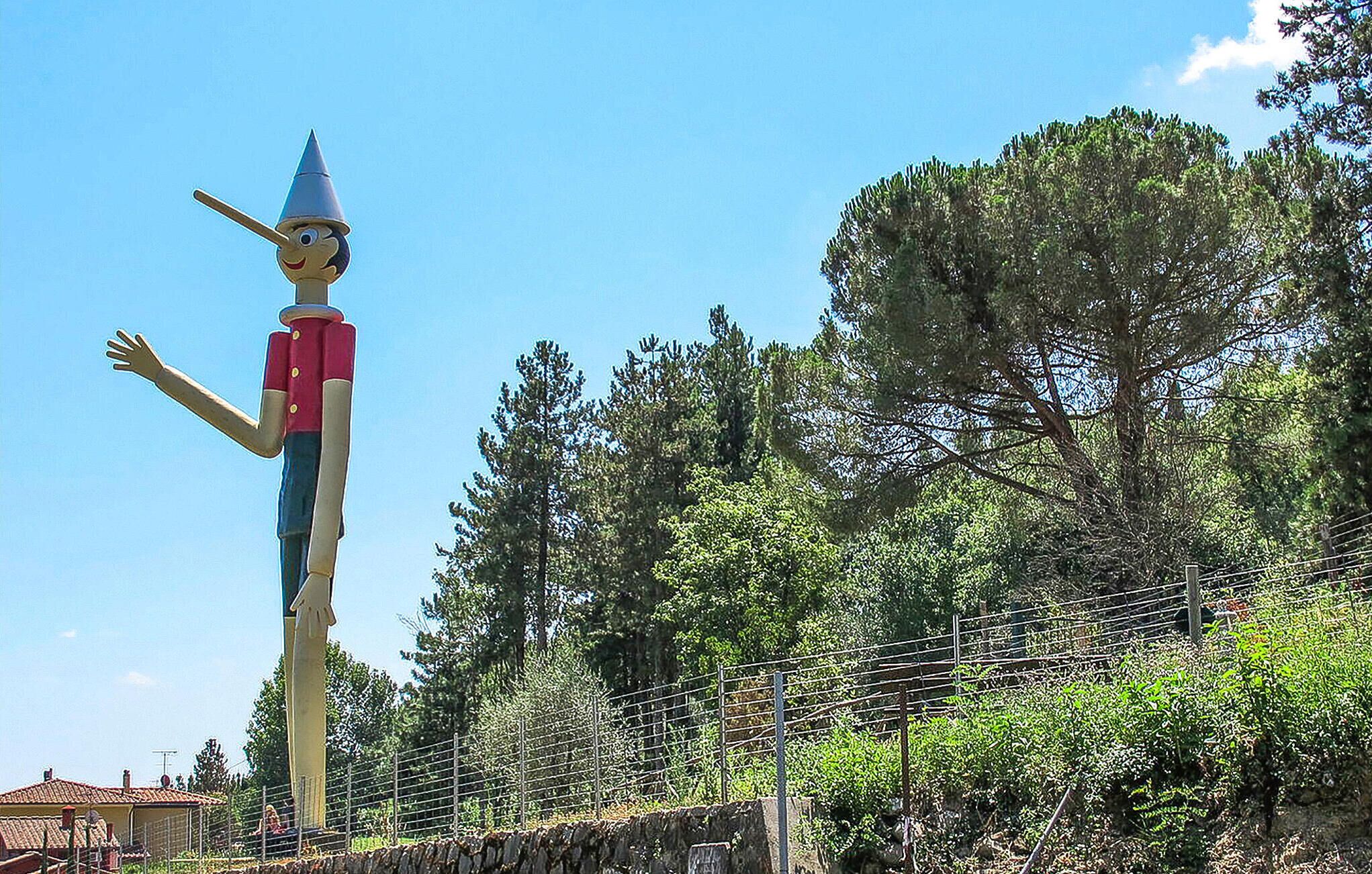 A tall, colorful statue of a character with a pointed hat and outstretched arms, set against a backdrop of trees and a stone wall.