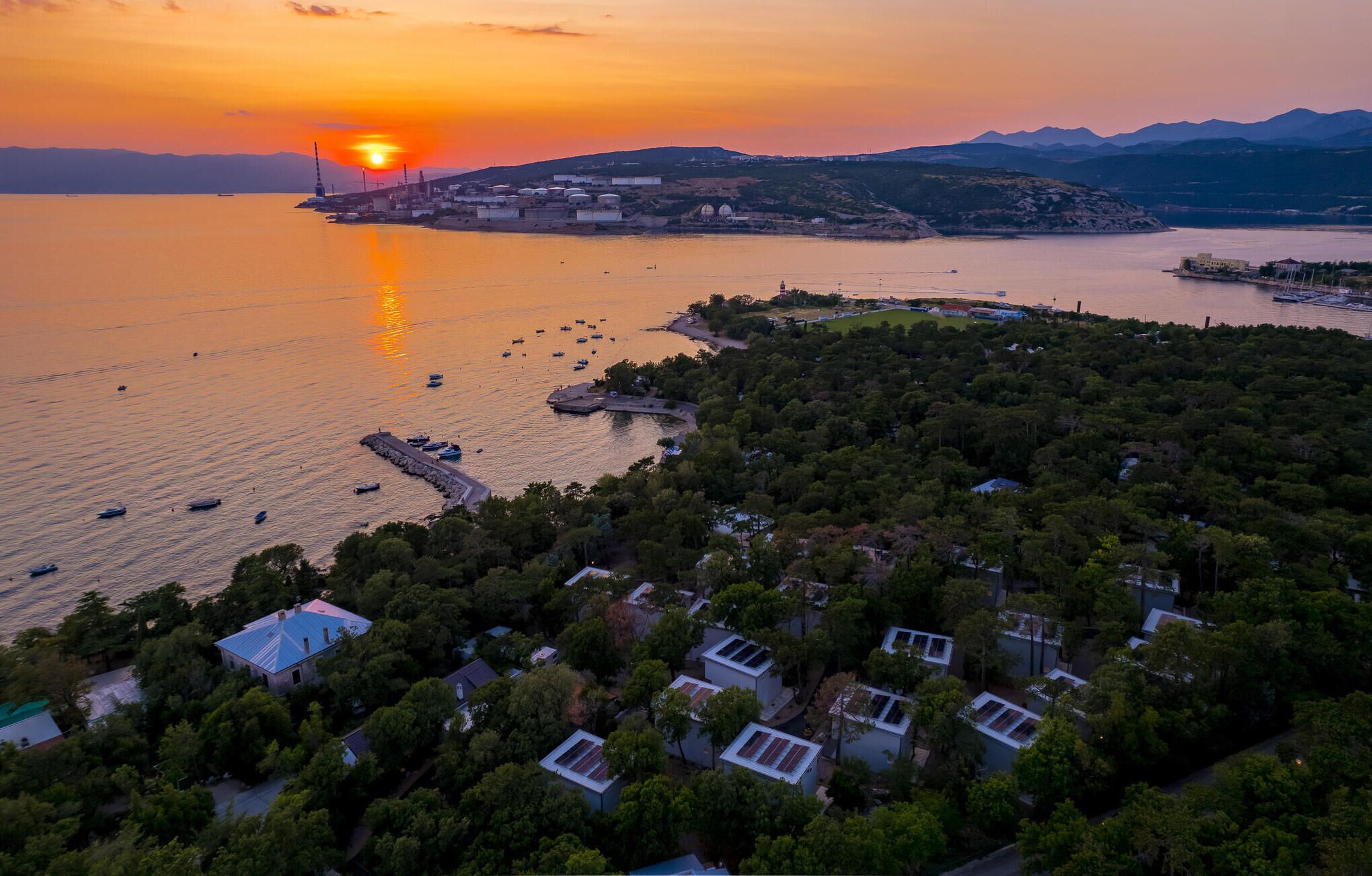 Vista aèria d'una zona costanera amb edificis residencials, un port esportiu i un atardeciment sobre l'aigua.