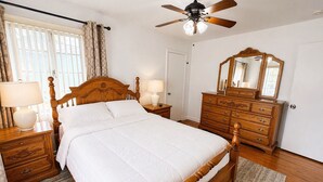 Un dormitorio con una cama de madera, una mesita de noche con lámpara, un aparador con espejo y un ventilador de techo.