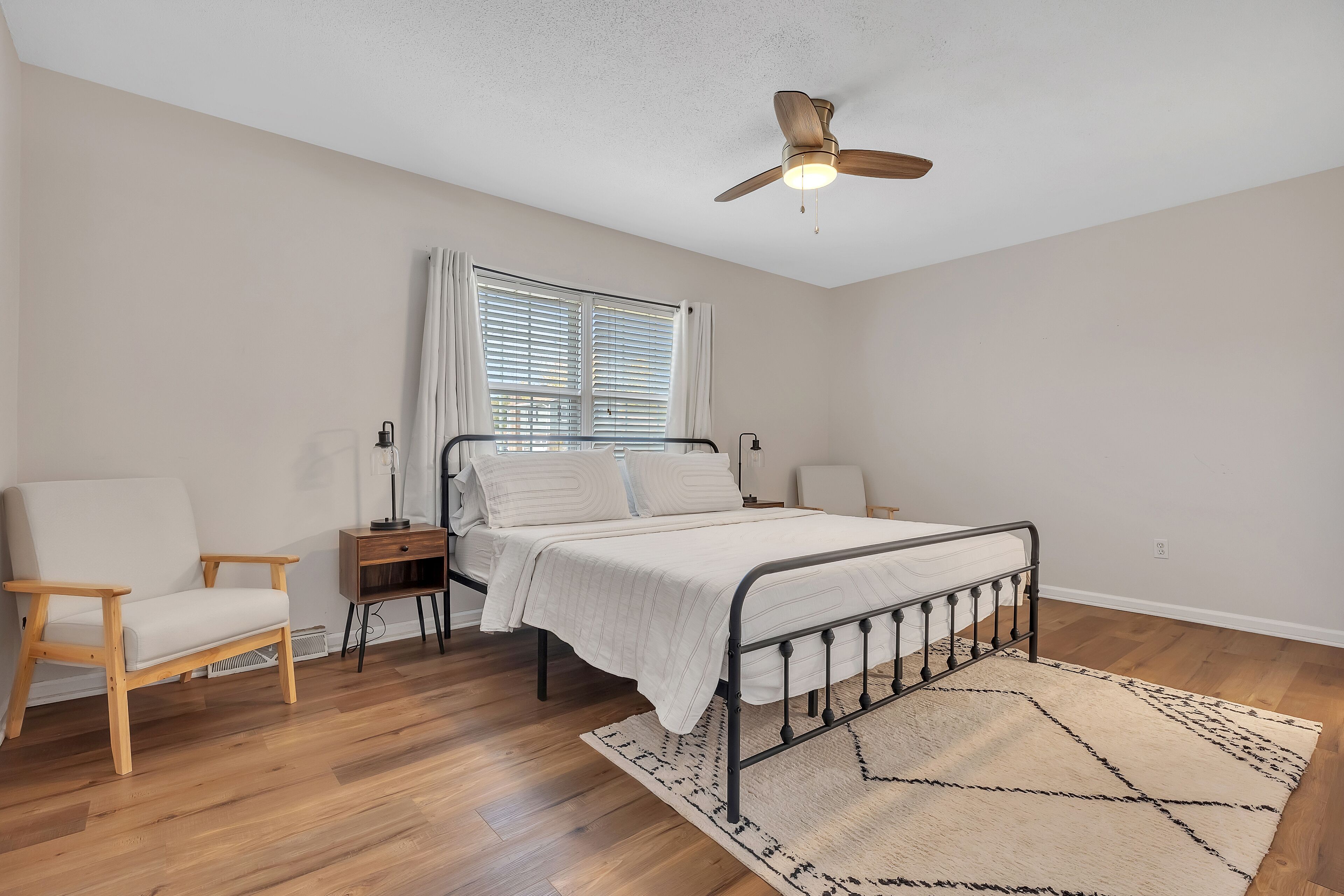 A bedroom with a bed, a chair, a nightstand, a ceiling fan, and a window with blinds.