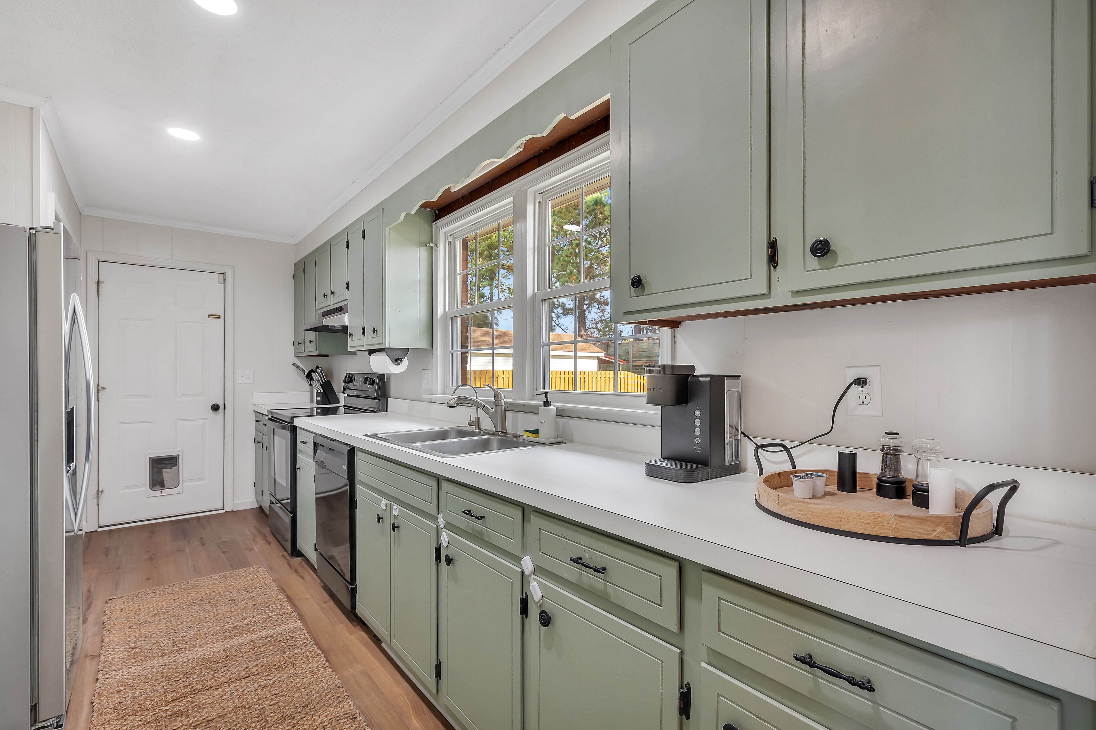 A kitchen with green cabinets, a white countertop, a coffee maker, and a window with a view of a fence.