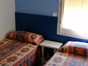 Une chambre avec deux lits, chacun agrémenté de draps à motifs, une petite table blanche et une fenêtre avec des stores.