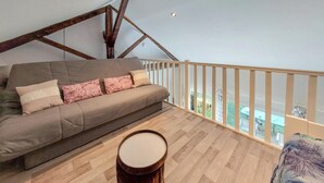 A loft-style living room with a wooden floor, a grey sofa with floral cushions, a drum, and a balcony with a railing.