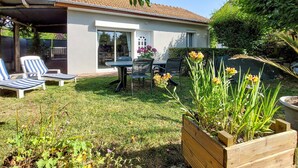 A house with a patio area featuring a table and chairs, surrounded by a garden with various plants and flowers.