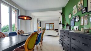 A modern kitchen with a dining area, featuring a large window, a hanging light fixture, and a green accent wall with various decorations. - Beautiful home in étretat with WiFi (Étretat)