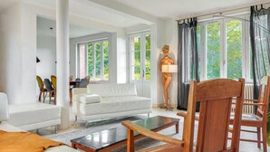 A modern living room with a white sofa, a glass coffee table, and wooden chairs. There are large windows with curtains, a lamp, and a statue. - Beautiful home in étretat with WiFi (Étretat)