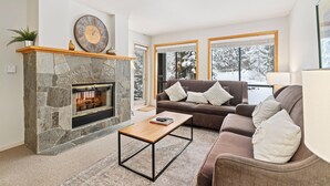 A living room with a stone fireplace, a brown sofa, a wooden coffee table, and large windows with a snowy view. - Powderhorn by Whistler Premier (Whistler)