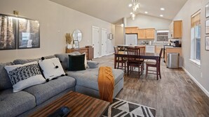 A modern living room with a grey sectional sofa, wooden coffee table, and a dining area with a wooden table and chairs. A kitchen with wooden cabinets and a refrigerator is visible in the background.