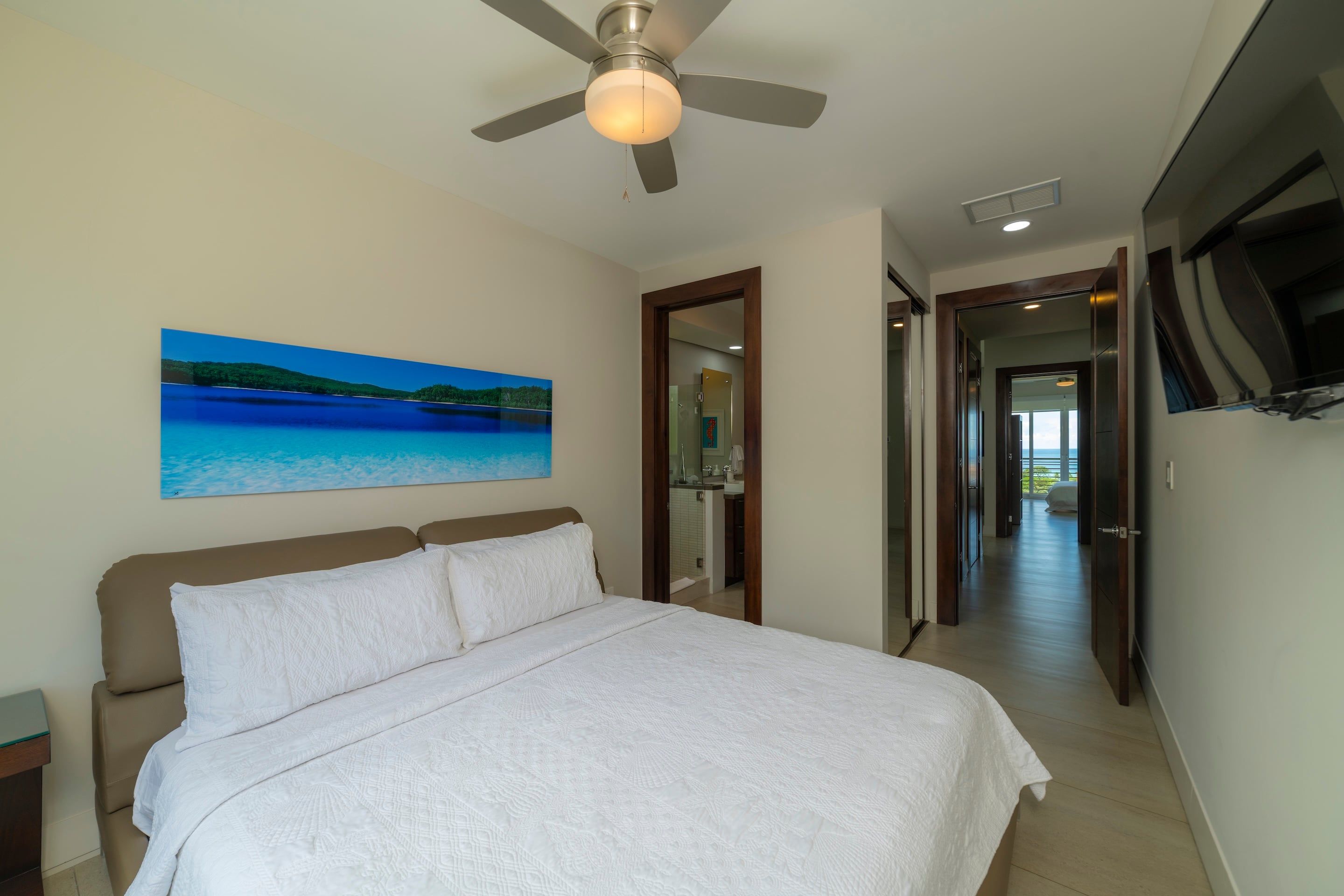 A bedroom with a bed, a ceiling fan, a painting of a beach, and a hallway leading to a bathroom.