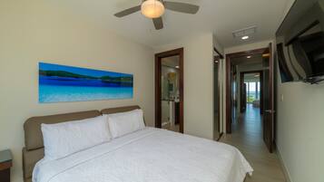 A bedroom with a bed, a ceiling fan, a painting of a beach, and a hallway leading to a bathroom.