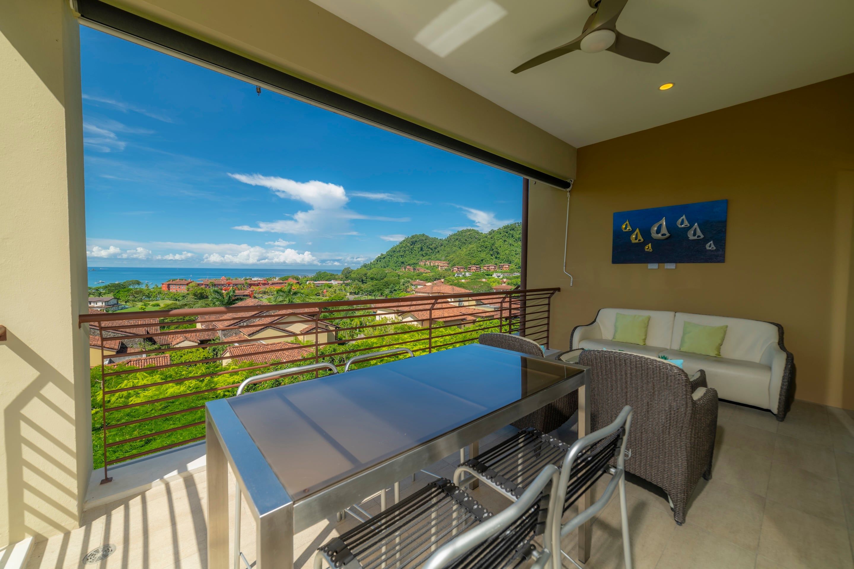 Ein Balkon mit Glastisch und Stühlen, einem weißen Sofa und einem Deckenventilator.