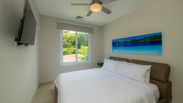 A bedroom with a bed, a ceiling fan, a TV mounted on the wall, a window with a view of greenery, and a painting on the wall.