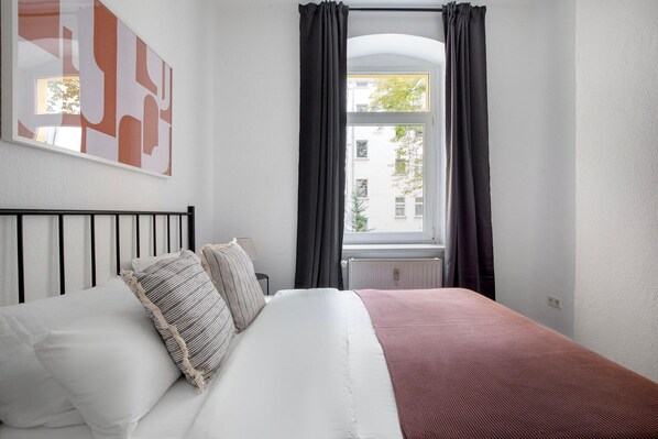 Un dormitorio con cama, ventana con cortinas, radiador y un cuadro en la pared.