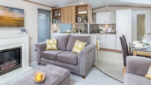 A modern living room with a fireplace, grey sofa, ottoman, and dining area with a table and chairs. - Autograph Junior Lodge 2 Bed with Hot Tub (Ormside, Appleby-in-Westmorland)