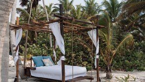 A wooden bed with white bedding and blue pillows, set on a sandy beach with palm trees in the background. - Playa Sonrisa Hotel (Xcalak)