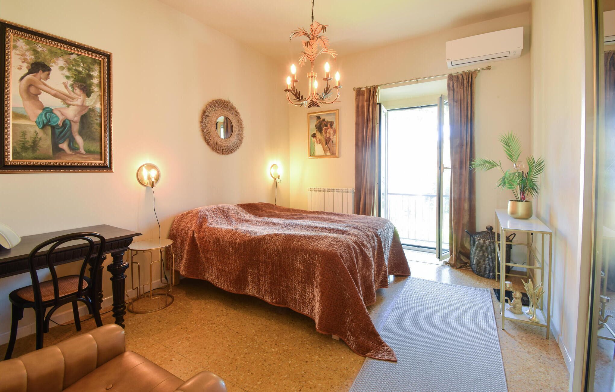 A bedroom with a large bed, a desk with a chair, a mirror, a chandelier, and a potted plant.