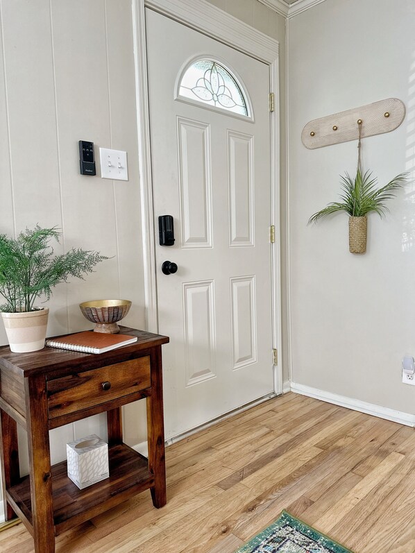 A wooden table with a potted plant and a bowl, a white door with a glass panel, a wall-mounted light fixture, and a small rug on the floor.