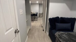A narrow hallway with a door on the left, a dark-colored sofa on the right, and a dining area in the background. - Cozy basement/studio apartment with kitchenette and bathroom! (Boston)