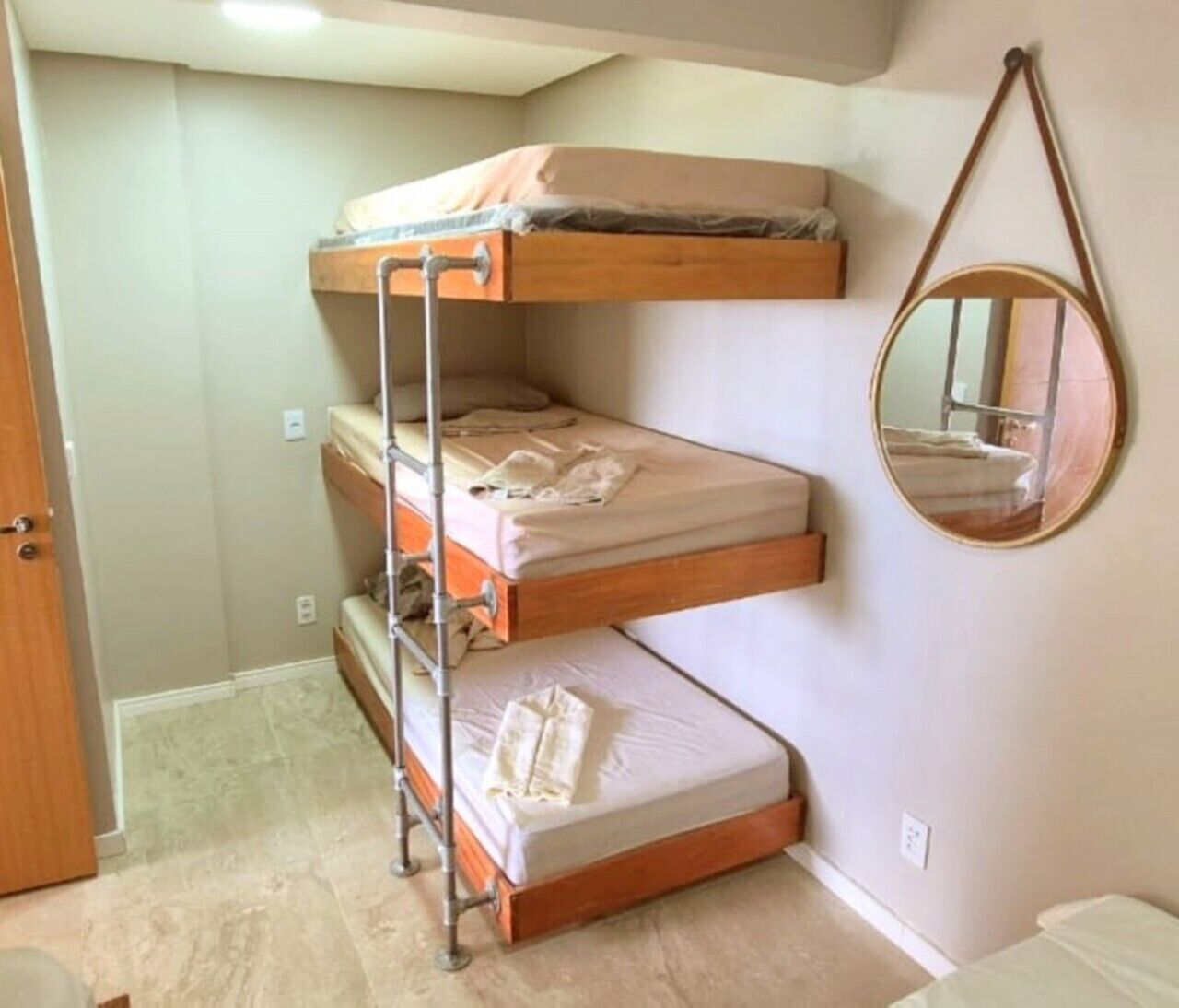 Una cama litera con escalera de metal, un espejo redondo en la pared y una puerta a la izquierda.