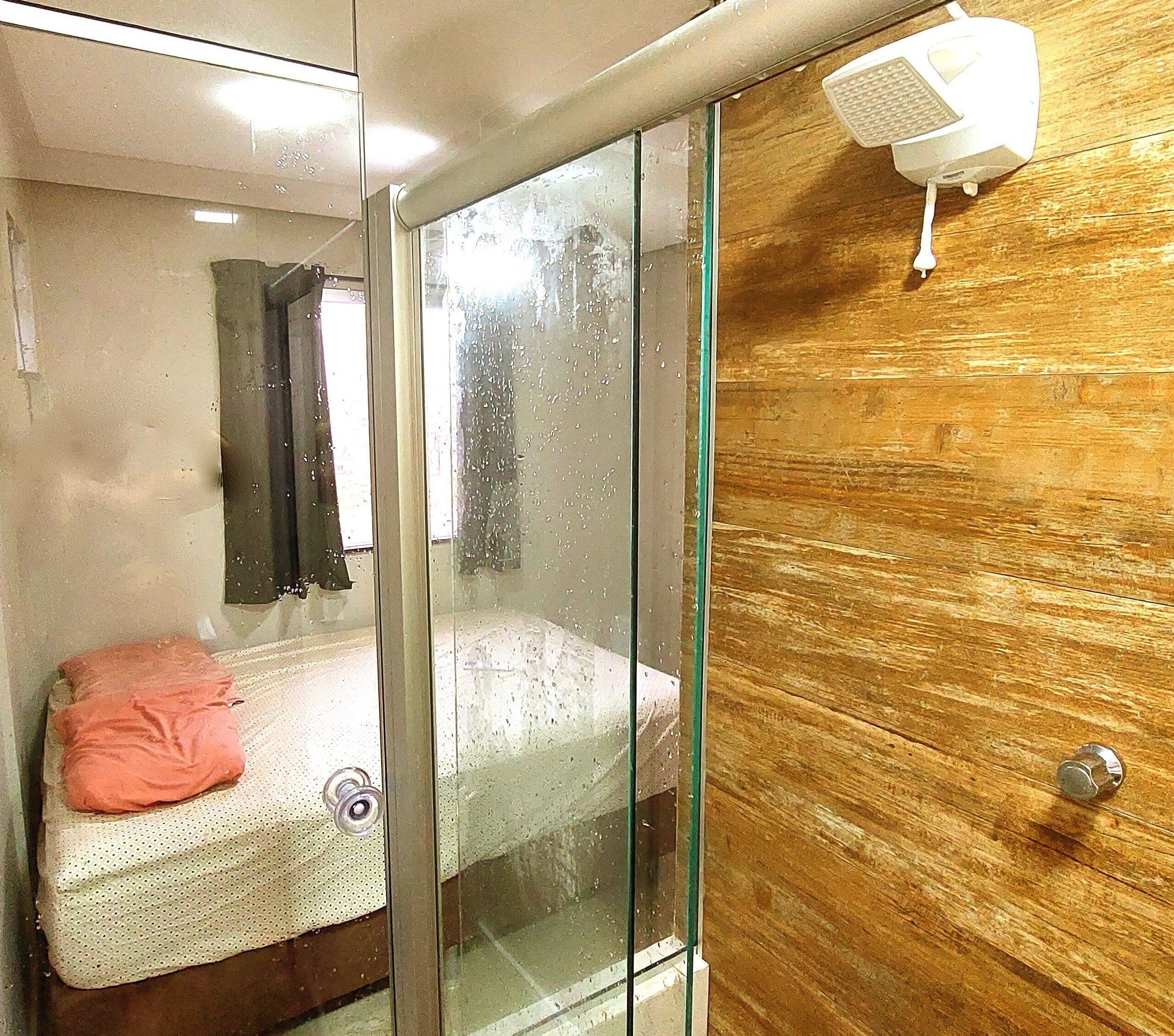 A narrow, glass-enclosed room with a bed, a wooden door, and a small white device on the wall.