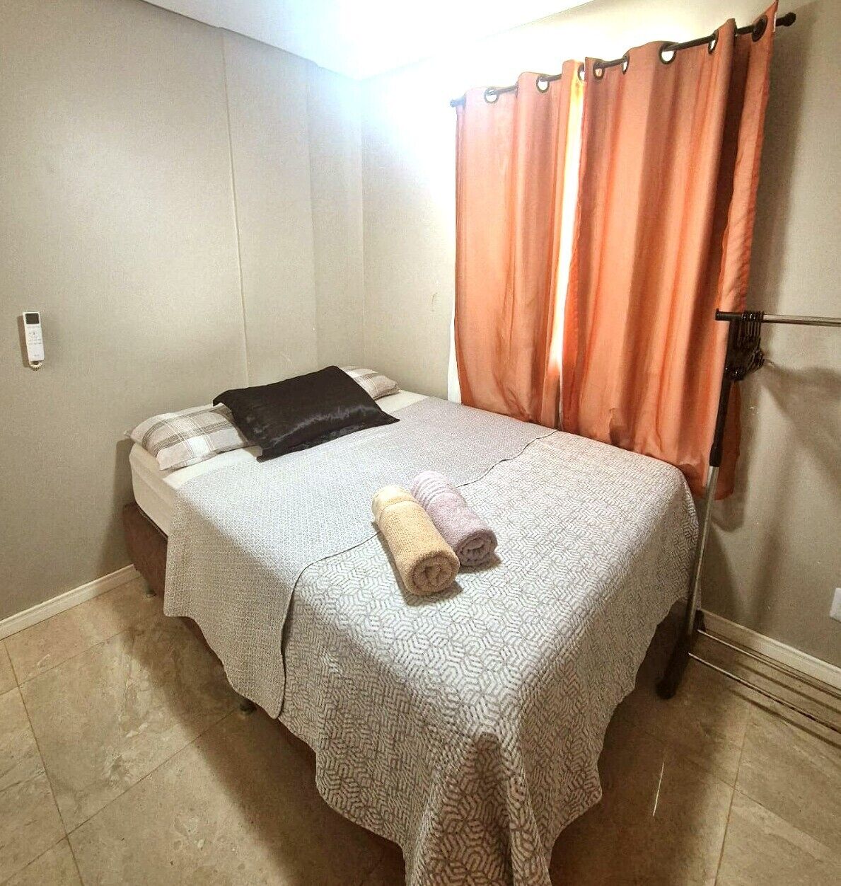 Una cama individual con una colcha estampada, dos toallas enrolladas, un almohán negro y una cortina naranja.