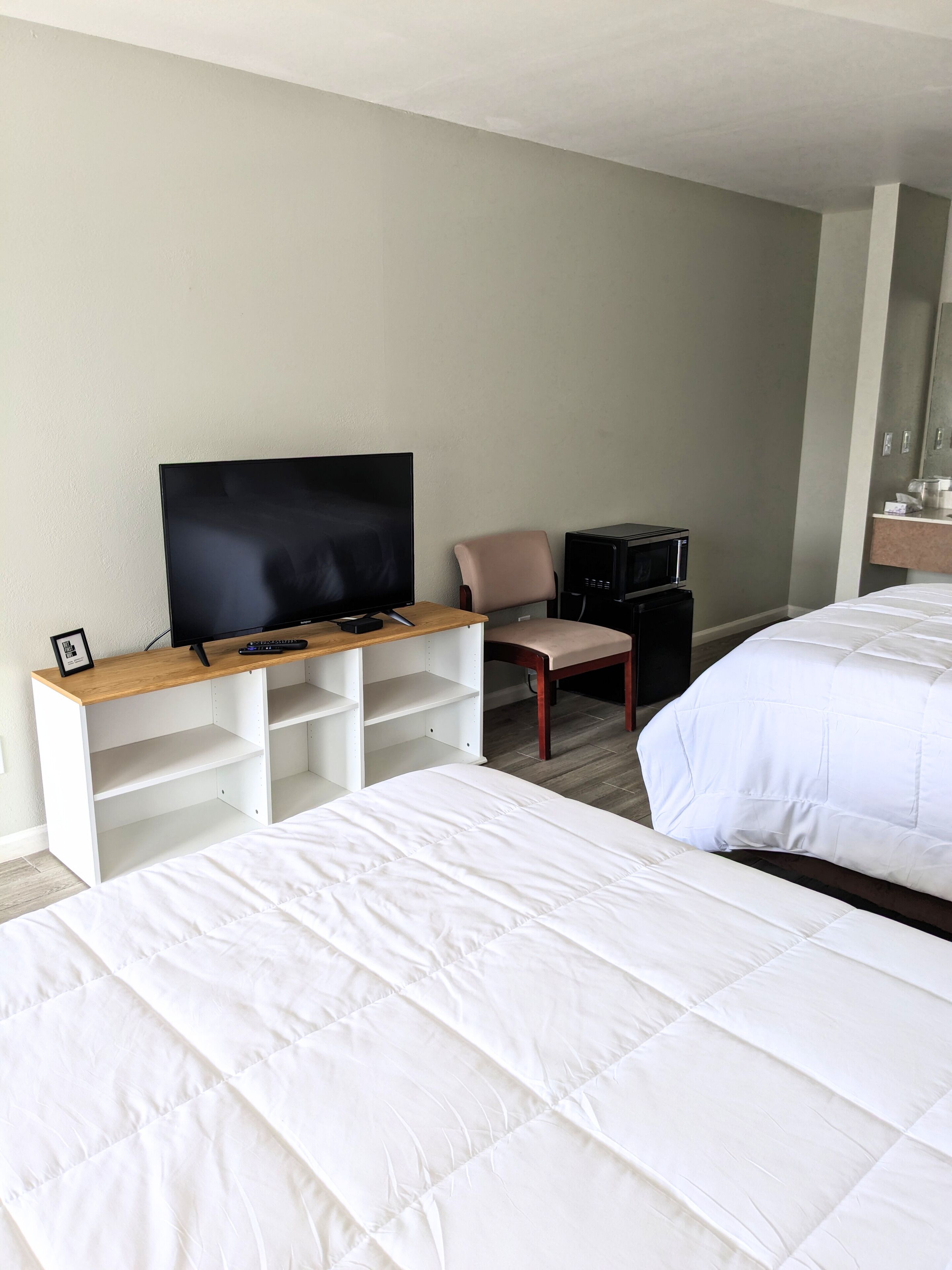 A hotel room with a bed, a TV on a wooden stand, a chair, a desk with a computer, and a microwave.