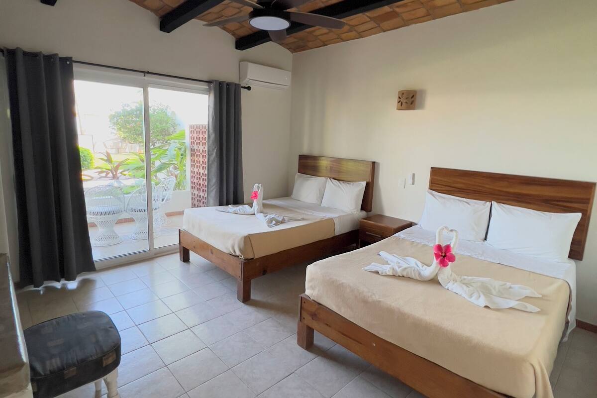 A hotel room with two wooden beds, a ceiling fan, and a view of a garden through a sliding glass door.