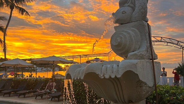 Una piscina con una fuente decorativa y una puesta de sol al fondo.