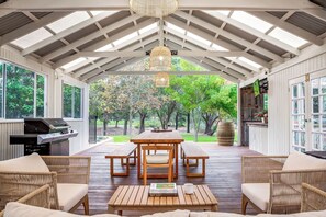 Eine überdachte Terrasse im Freien mit Holztisch und -stühlen, einem Grill und Korbmöbeln.
