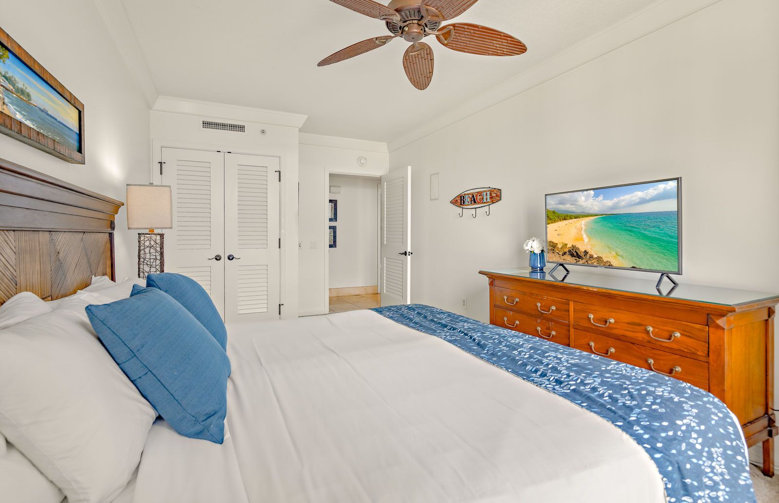 Ein Schlafzimmer mit einem großen Bett, einer Holzkommode, einem Deckenventilator und einem Fernseher, der eine Strandansicht zeigt.