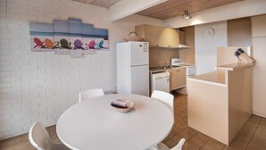 A compact kitchen with white appliances, a dining area with a round table, and a beach-themed wall art. - Pacific Heights - Standard Apartment (Merimbula)