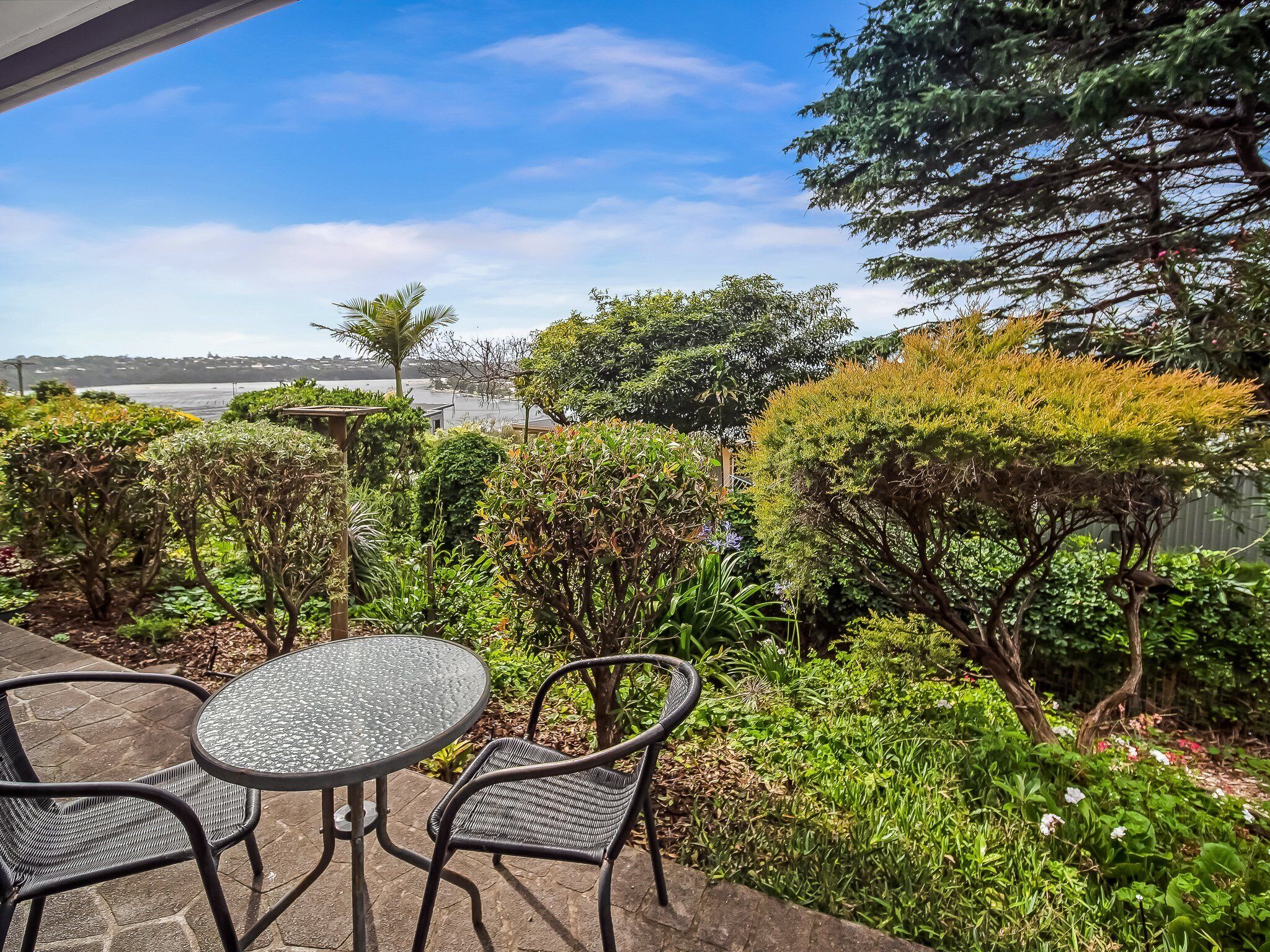 A patio with a round table and two chairs, surrounded by lush greenery and a view of a body of water.