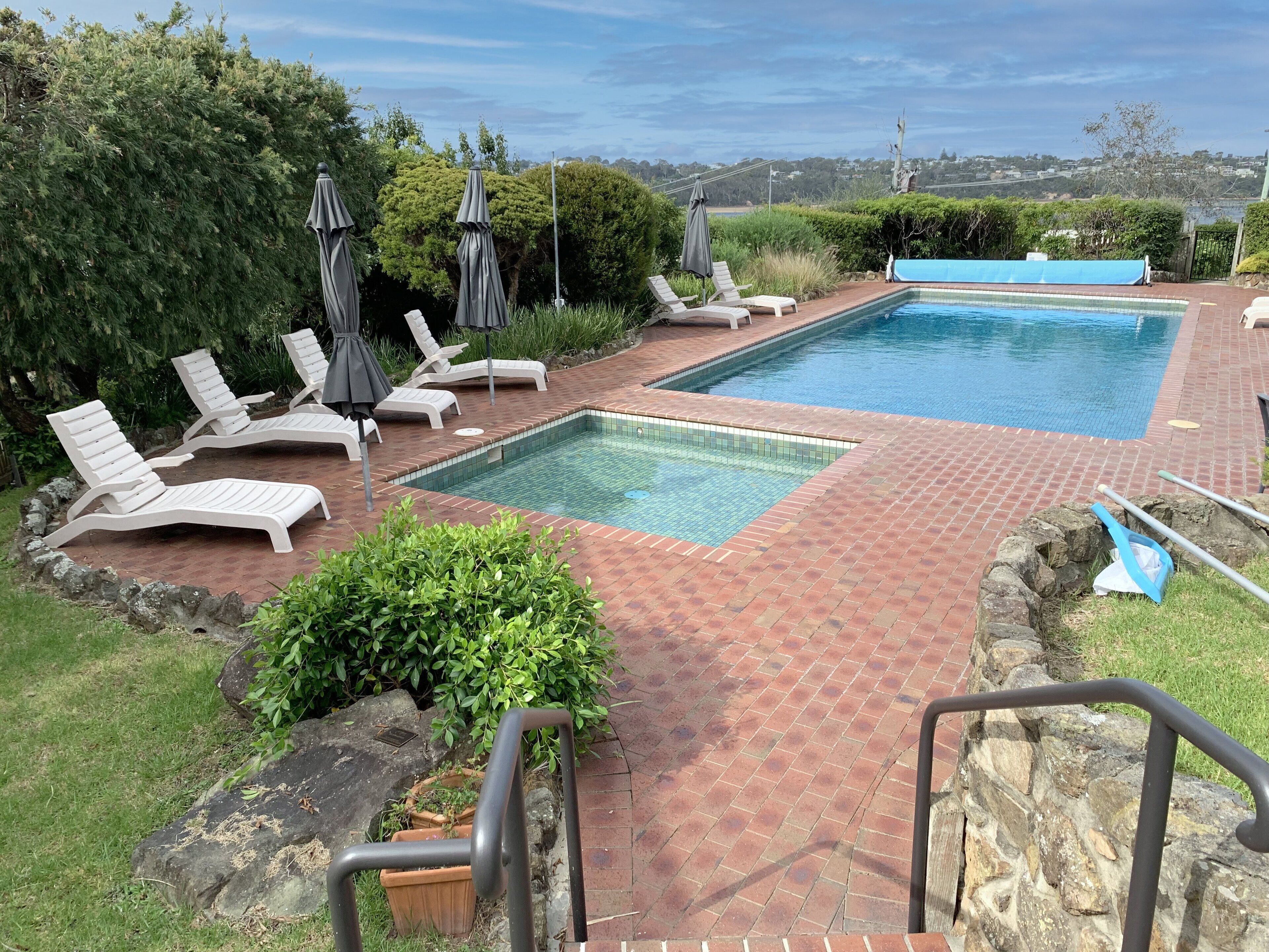 Une piscine rectangulaire divisée en deux zones distinctes, entourée d’une terrasse en briques. On y trouve des chaises longues blanches et des parasols, et la vue s’étend sur un paysage lointain.