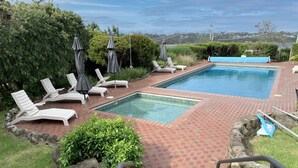 A rectangular pool with two separate sections, surrounded by a brick patio. There are white lounge chairs and umbrellas, and a view of a distant landscape. - Pacific Heights - Standard Apartment (Merimbula)
