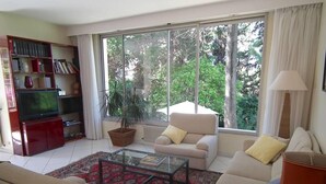 A living room with a sofa, armchair, coffee table, bookshelf, TV, and a large window with a view of trees.