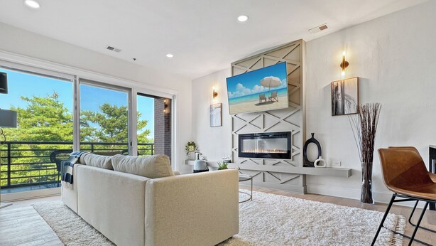 A modern living room with a fireplace, a large TV, and a balcony with a view of trees and sky. - Luxury 2Bed w/ Amazing Views (Cornelius)