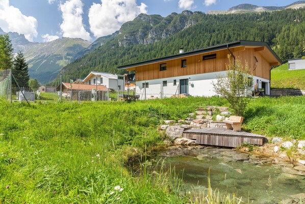 Un bâtiment moderne doté d’une façade en bois, d’un petit quai et d’un ruisseau aux eaux claires, situé dans une région montagneuse.
