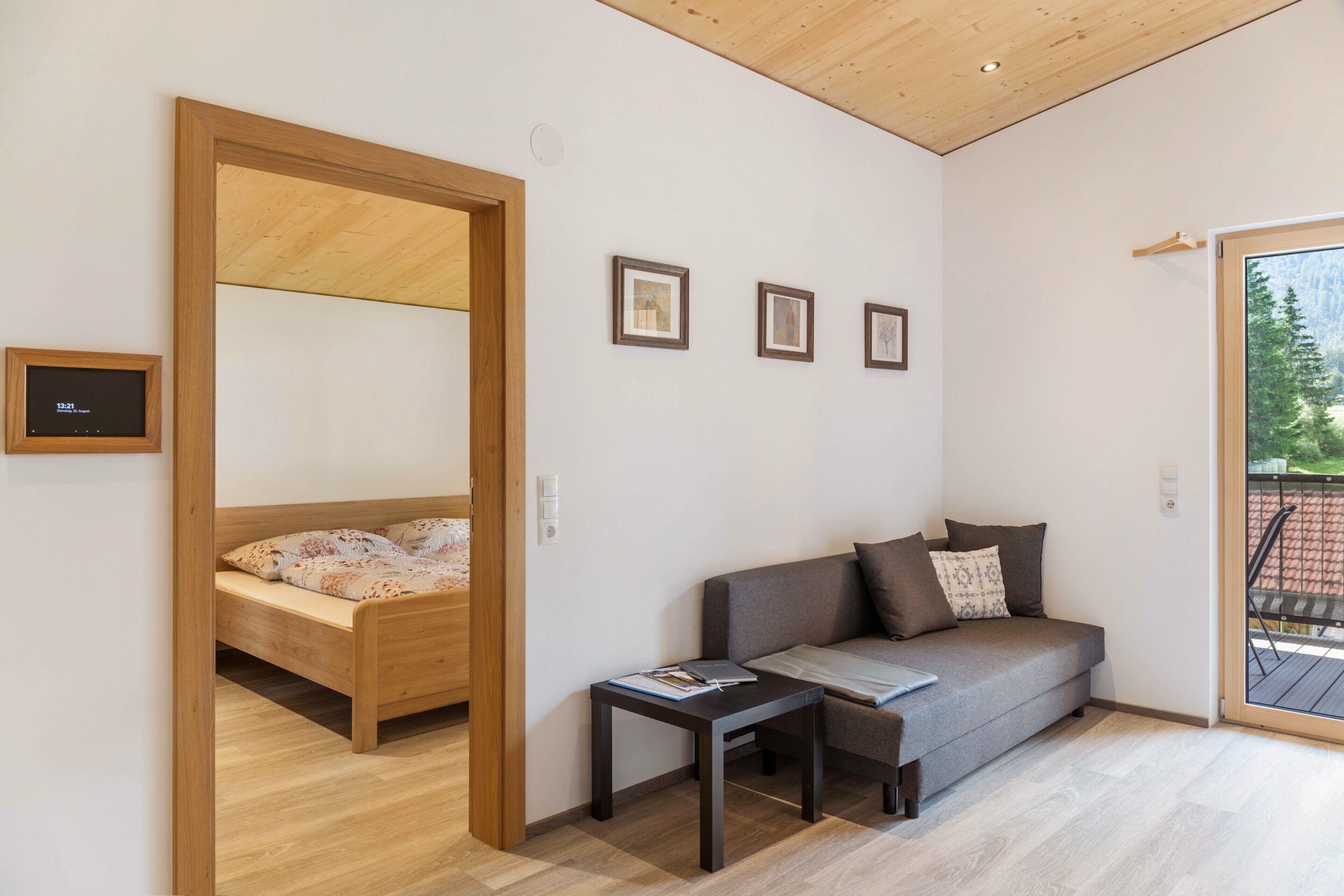 A room with a bed, a sofa, a small table, and a door leading to another room. There are three framed pictures on the wall.