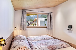 Une chambre avec un plafond en bois, une grande fenêtre ornée de rideaux à motifs, un lit avec une literie à fleurs et une prise de courant fixée au mur.