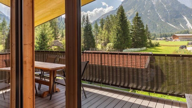 Eine Holzdiele mit Tisch und Stühlen, mit Blick auf eine Berglandschaft mit Bäumen und einem Gebäude.