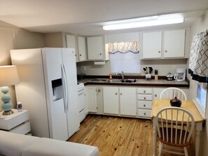 A compact kitchen with white cabinets, a refrigerator, a stove, and a sink. There is a dining table with a chair, a lamp, and a window with blinds. - Island Paradise with private beach & private bay sunset views. (Key Largo)