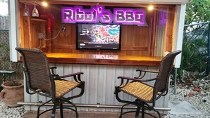 A wooden bar area with two wicker chairs and a neon sign reading "Rib's BBQ". - Island Paradise with private beach & private bay sunset views. (Key Largo)