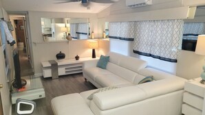 A modern living room with a white sofa, a ceiling fan, and a large window with patterned curtains. - Island Paradise with private beach & private bay sunset views. (Key Largo)