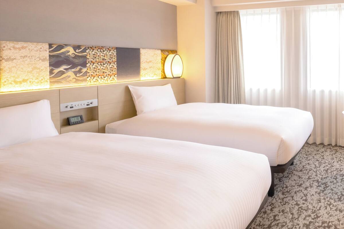 A hotel room with two beds, a headboard with a decorative panel, a bedside table with a lamp, and a window with curtains.