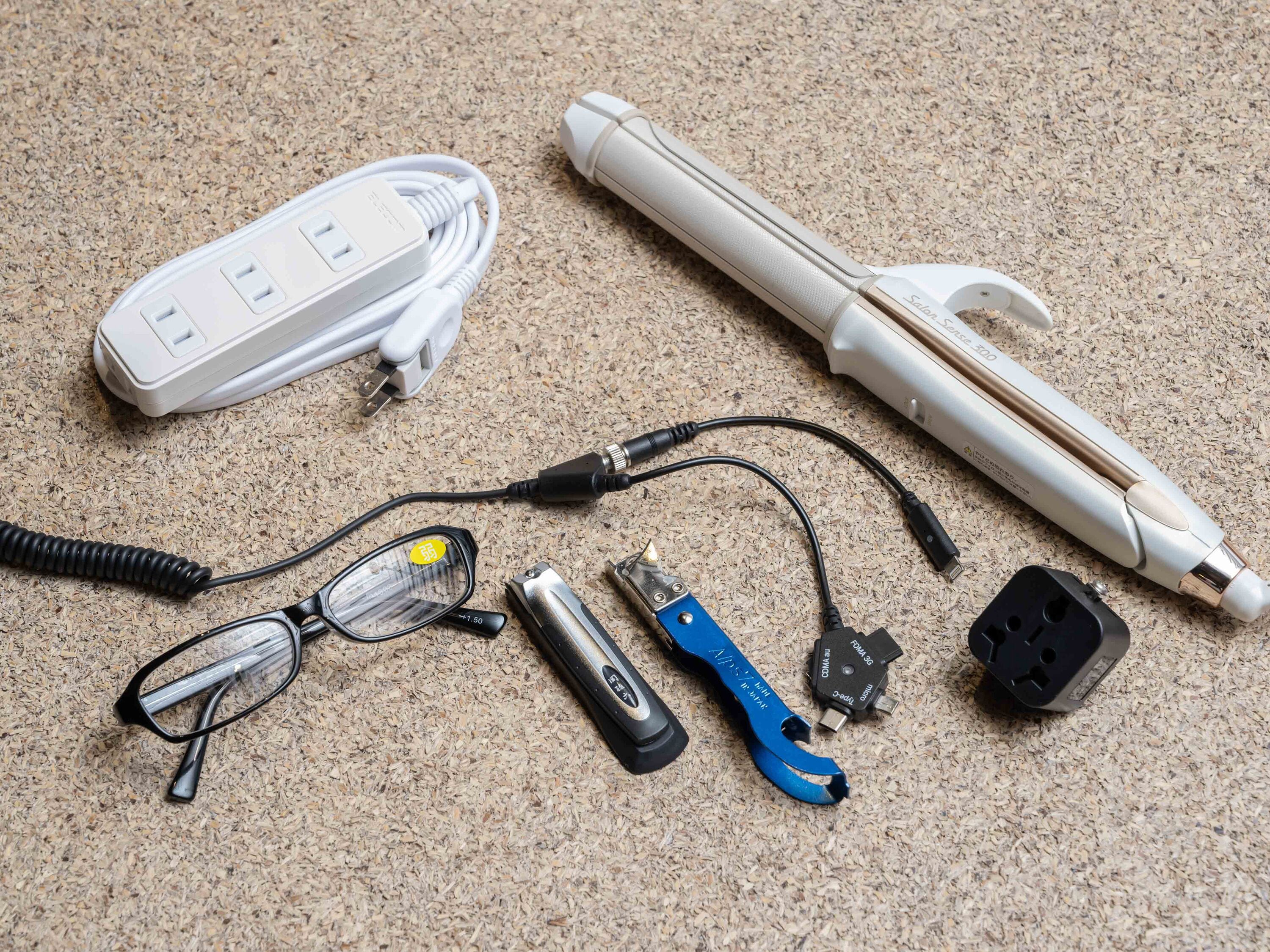 A power strip, a white cylindrical device, a pair of glasses, a blue tool, and a black power adapter on a textured surface.