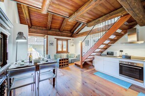 Une cuisine avec des poutres en bois, un coin repas avec des chaises, un escalier et une vue sur l’extérieur.