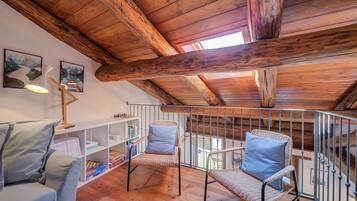 Une pièce mansardée chaleureuse avec des poutres en bois, un balcon doté d’une rambarde en métal, des fauteuils en osier avec des coussins et une bibliothèque.