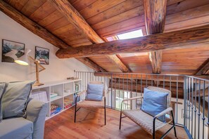 A cozy attic room with wooden beams, a balcony with a metal railing, wicker chairs with cushions, and a bookshelf.