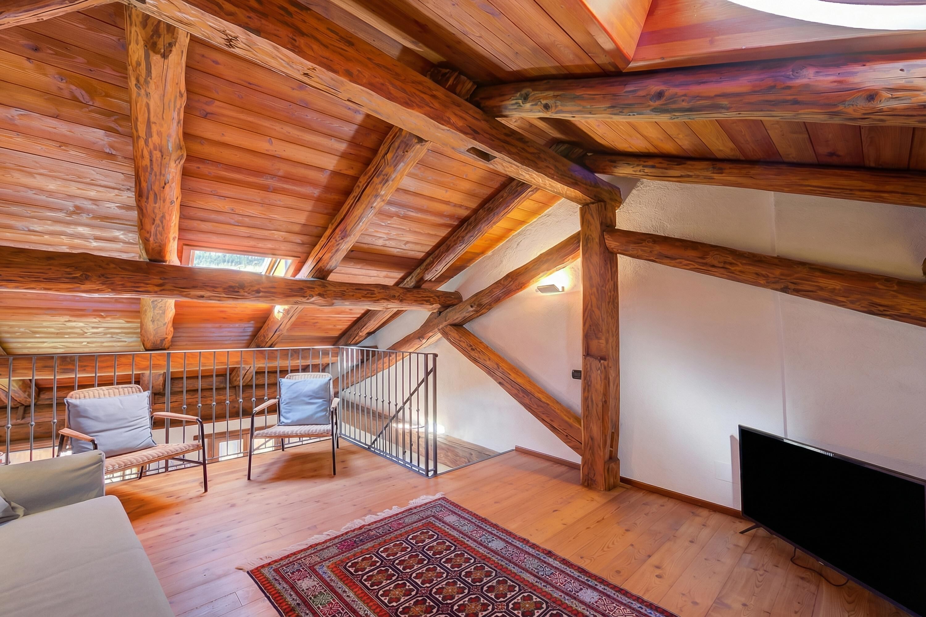 Una habitación amplia en el ático, con vigas de madera a la vista, un ventanal grande, un televisor de pantalla plana y una zona de estar acogedora con sillas y un sofá.
