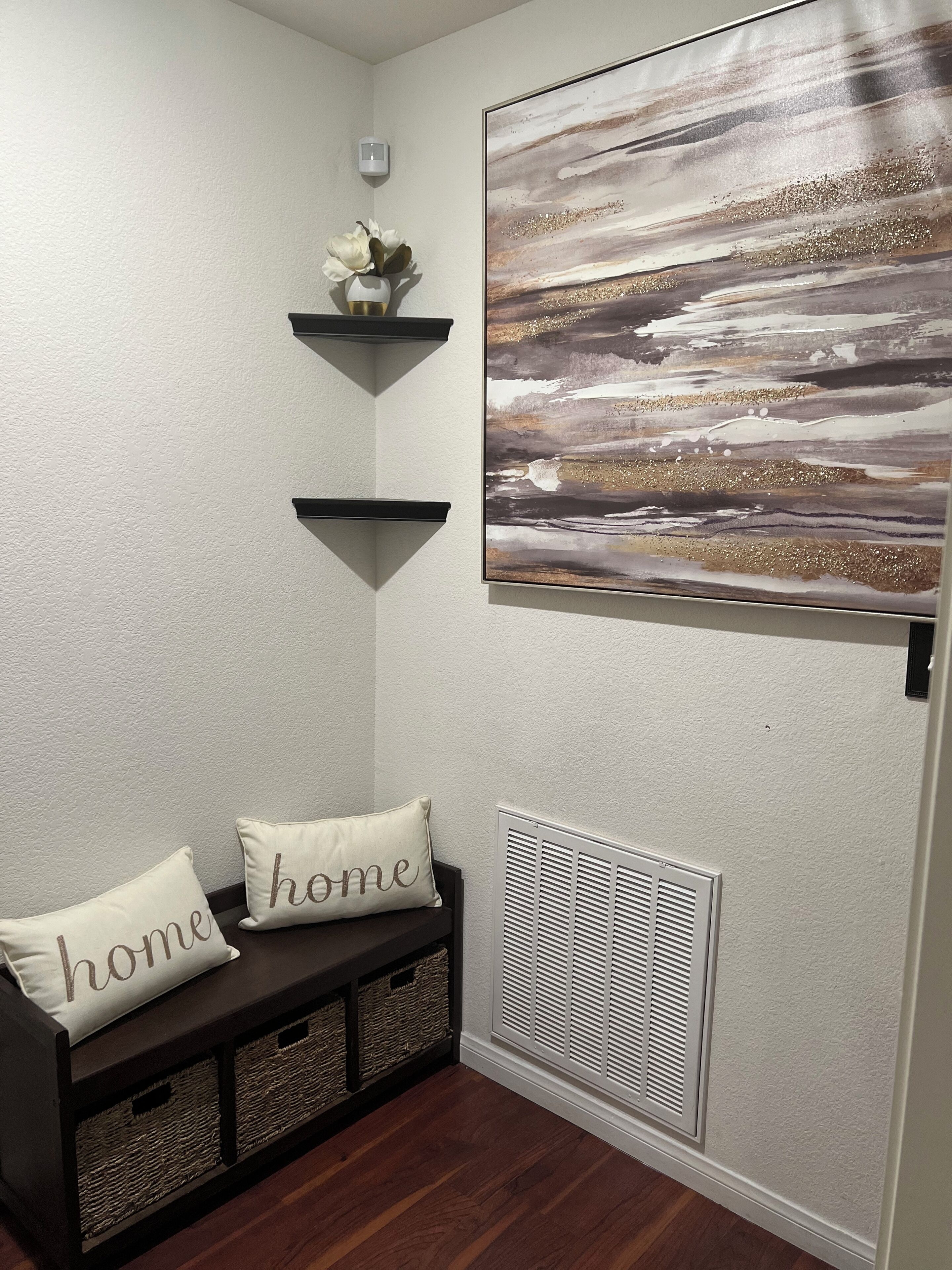 Ein Flur mit einer Bank, zwei Kissen mit dem Aufdruck ‚home‘, einem an der Wand befestigten Regal mit einer Vase und einem großen abstrakten Gemälde.